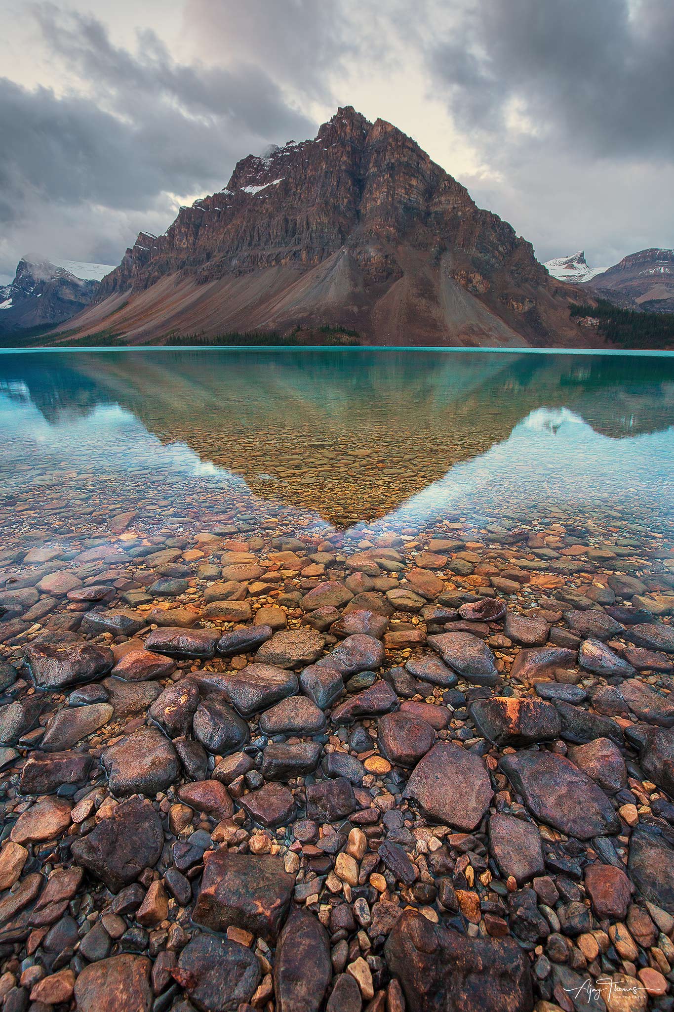 Majestic peaks of the mountain rising in the background, with the serene waters of the lake in the foreground, and the pebbles...